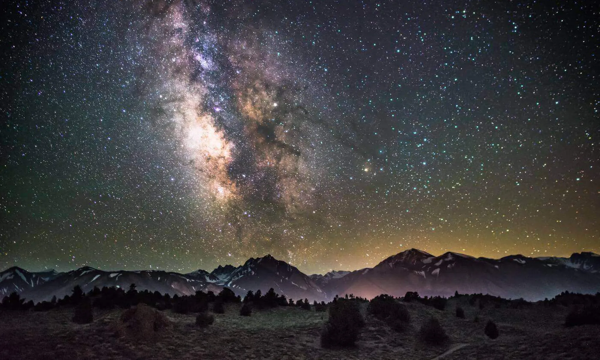 The milky way above a foggy landscape with mountains in the background and a forest in the foreground.